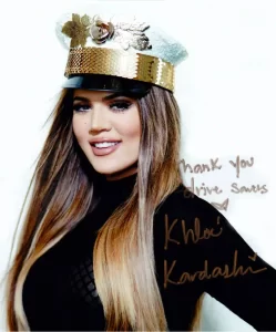 A woman with long hair wearing a decorative hat and a black outfit smiles. The image has handwritten text that says "Thank you drive safely" and is signed by "Khloé Kardashian.