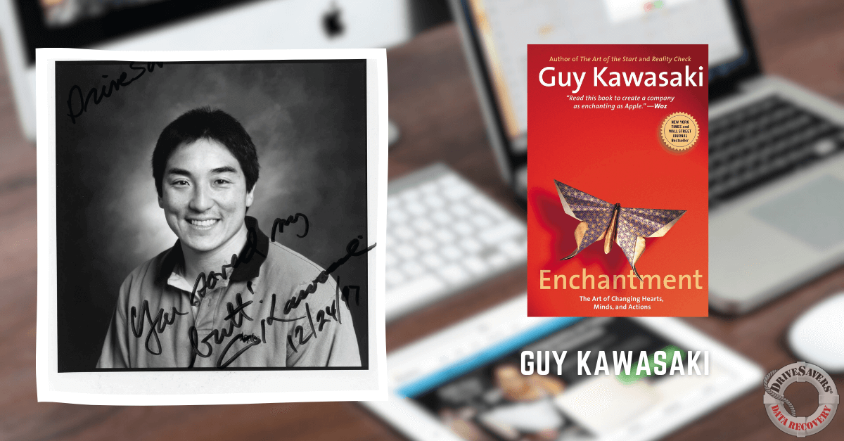 The image depicts a black and white photograph of a man who has autographed the photo. Next to him is a copy of "Enchantment" by Guy Kawasaki. In the background, you can see a laptop and computer screen that are intentionally blurred, drawing attention to the man and the book in focus.