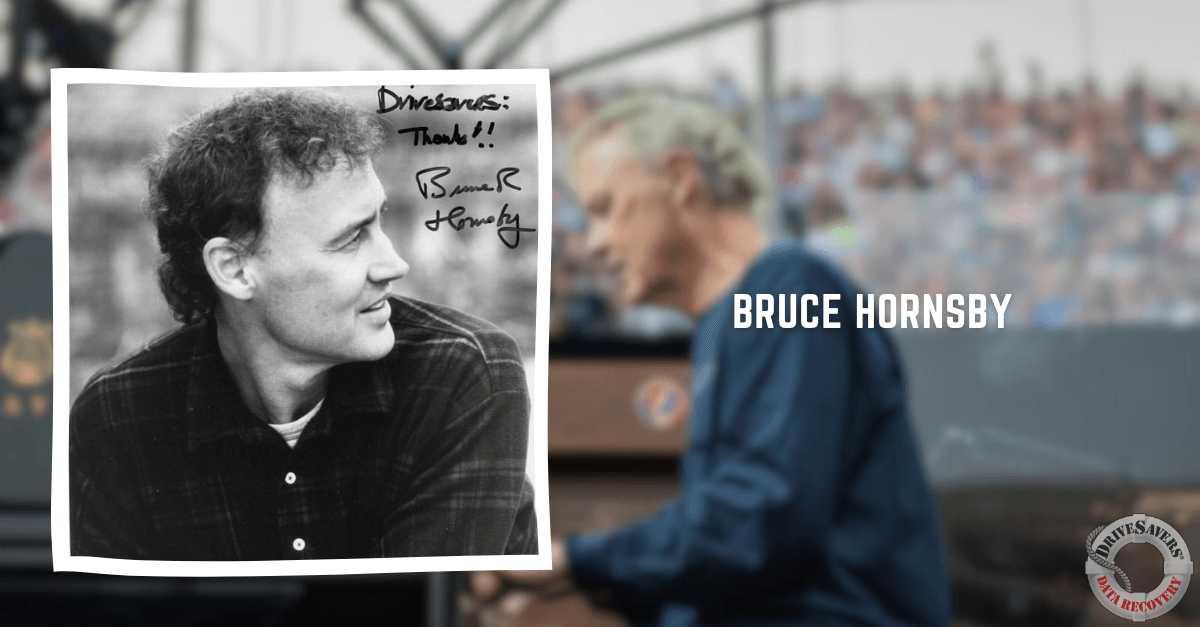 The black and white photograph captures a moment where a man with curly hair, wearing a plaid shirt, is gazing thoughtfully to the side. The image has been autographed with "Thanks!! Bruce Hornsby." In the background, slightly out of focus, another man is seated at a piano.