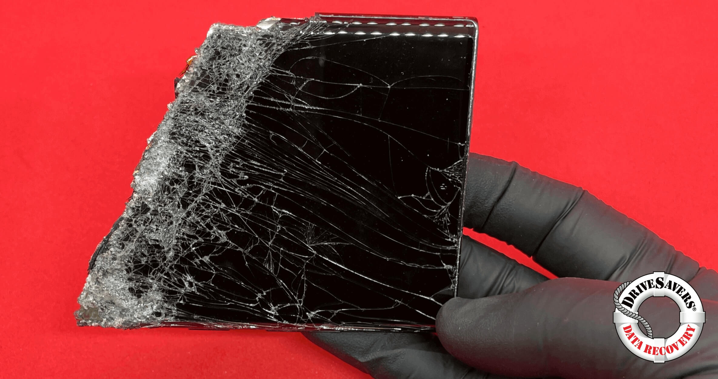 A gloved hand holds a shattered smartphone screen against a striking red background. The glass is cracked and splintered, revealing intricate fracture patterns. The Drivesavers Data Recovery logo sits confidently in the bottom right, promising to recover data from broken phones with precision and care.