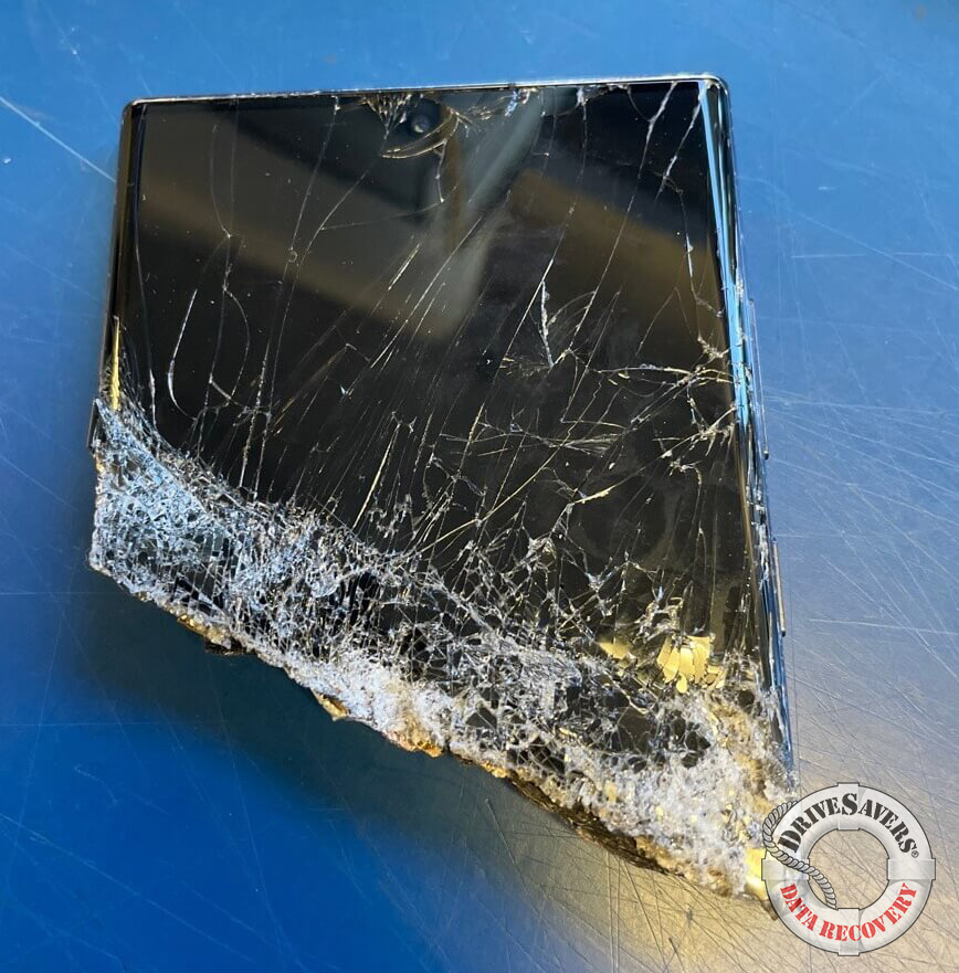 A shattered electronic device with severely cracked glass and missing pieces rests on a blue surface, silently pleading for rescue. A logo on the lower right corner reads "DRIVESAVERS DATA RECOVERY," hinting at hope to recover data from broken phone parts.