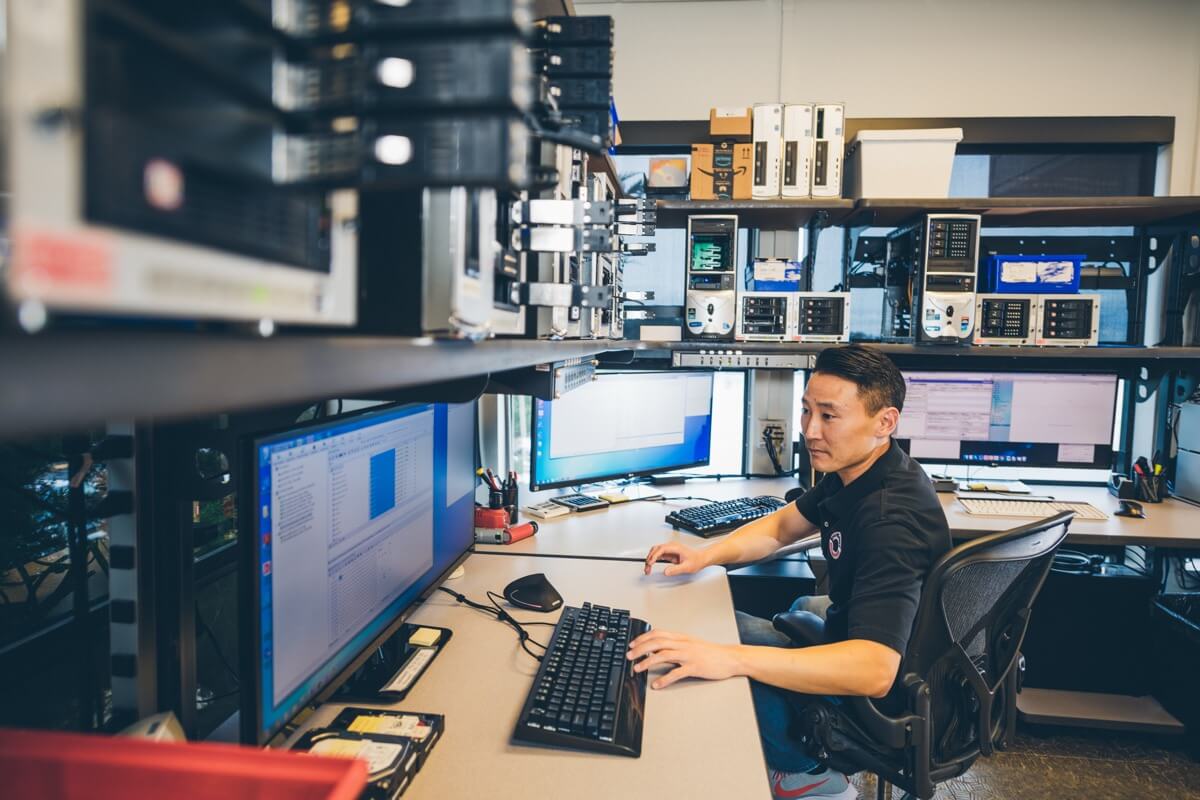 A person sits at a desk with multiple computer monitors displaying data and graphs. The room is filled with electronic equipment and shelving. The individual is focused on the screens, suggesting they are working or analyzing information.